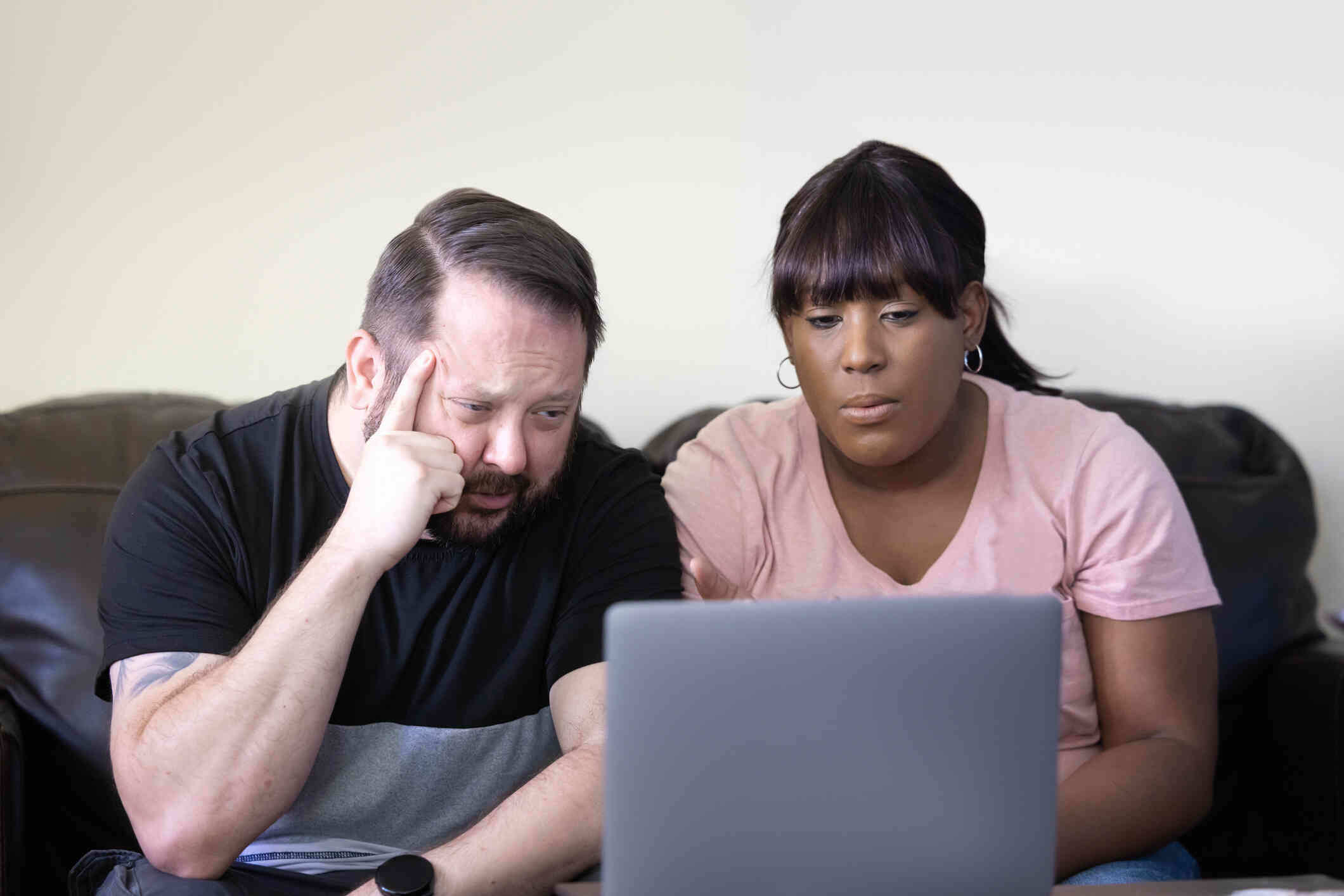 A couple seated on a couch appears worried as they look at the laptop screen together.